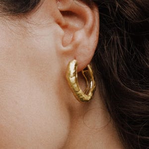 Puede incluir: Primer plano de un pendiente de aro dorado con un diseño texturizado y orgánico. El pendiente se lleva en una oreja con cabello castaño oscuro en el fondo. El pendiente tiene una forma irregular única.