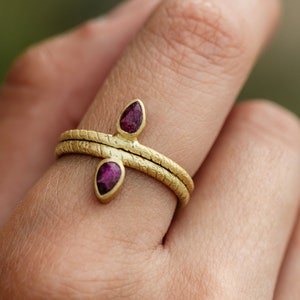 Könnte beinhalten: Ein goldener Ring mit zwei tropfenförmigen, violetten Edelsteinen, die in die Band gefasst sind. Der Ring ist texturiert und hat eine gehämmerte Oberfläche.