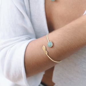 May include: A gold bracelet with a light blue gemstone. The bracelet is open and has a curved design. The gemstone is set in a gold setting.