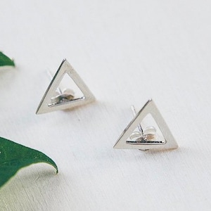 May include: A pair of silver triangle earrings with a hollow center. The earrings are on a white surface with green leaves.