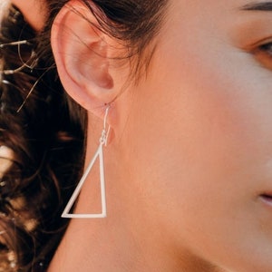 Könnte beinhalten: Eine Frau mit silbernen Dreiecksohrringen. Die Ohrringe sind schlicht und geometrisch.