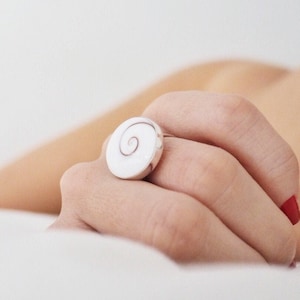 Puede incluir: Un anillo blanco con un diseño en espiral en un dedo con esmalte de uñas rojo.