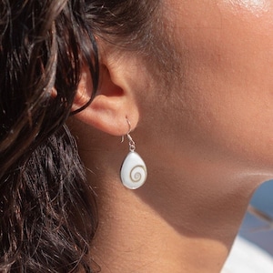 May include: A woman with long, dark brown hair wearing a white shell earring. The earring is a teardrop shape with a white shell and a silver swirl design.