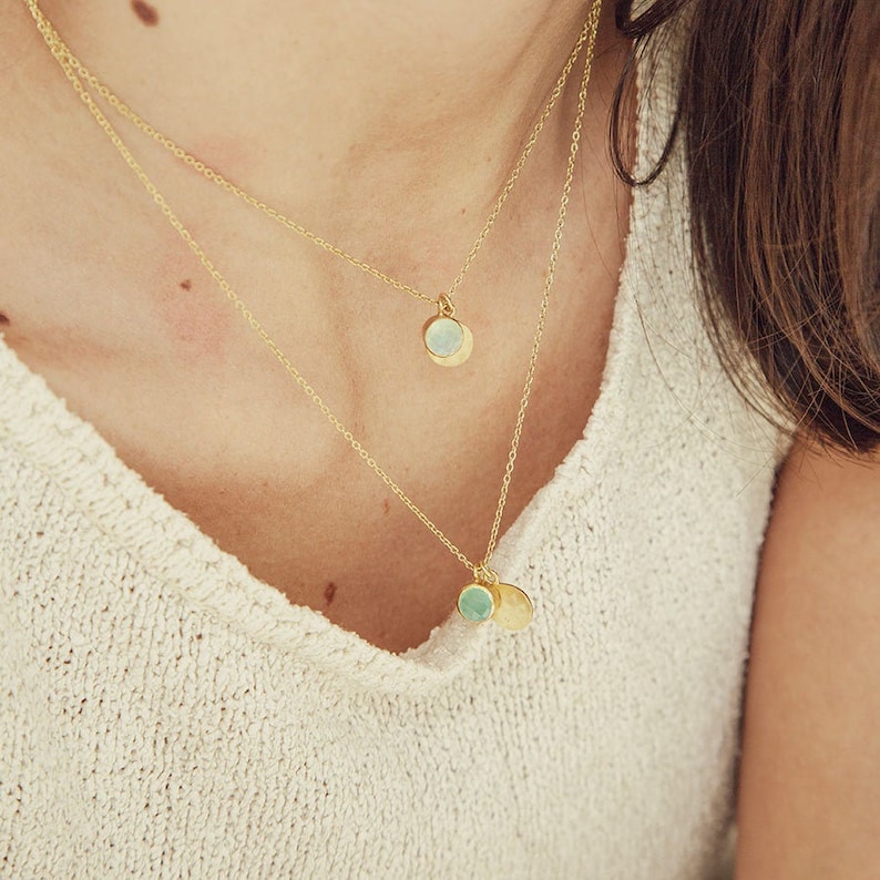 May include: Two gold necklaces with delicate chains and small round pendants. The top pendant is white and the bottom pendant is green.