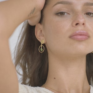 May include: Gold earrings with a small green stone pendant hanging from a circle. The earrings are worn by a person with brown hair.