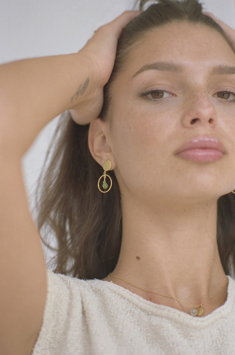 May include: Gold earrings with a small green stone pendant hanging from a circle. The earrings are worn by a person with brown hair.