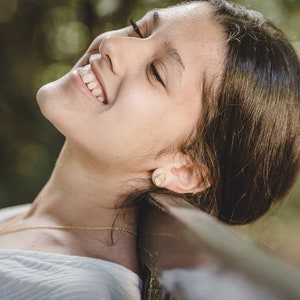 May include: A young person with long brown hair is lying on their back with their eyes closed and smiling. They are wearing a white shirt and a gold necklace with a small green stone pendant.