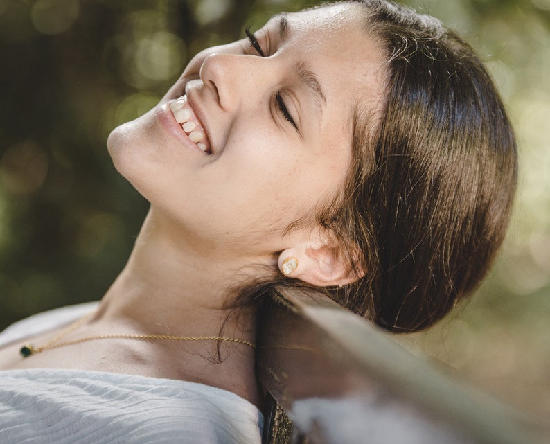 May include: A young person with long brown hair is lying on their back with their eyes closed and smiling. They are wearing a white shirt and a gold necklace with a small green stone pendant.