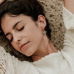 May include: A woman with long dark hair wearing a white sweater with a delicate gold and light blue flower earring.