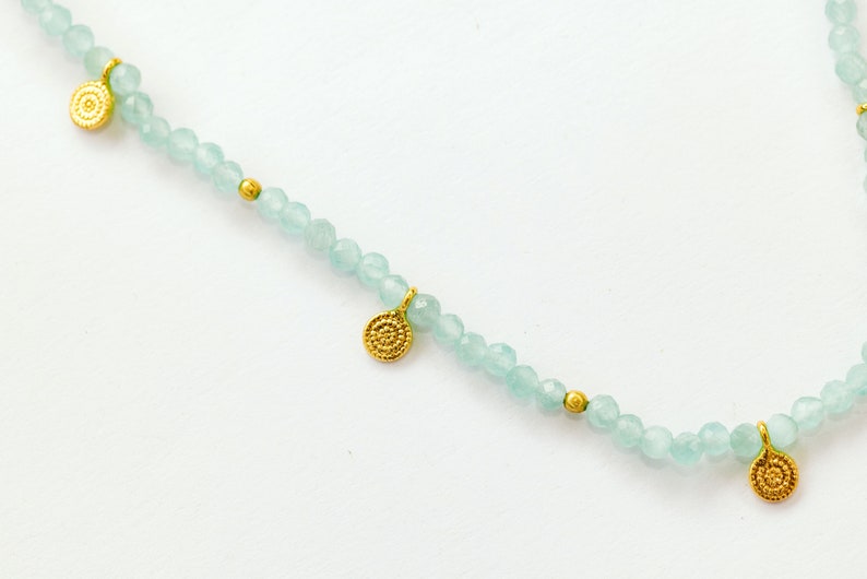 K&ouml;nnte beinhalten: Eine zarte Halskette mit hellblauen Perlen und goldenen Anh&auml;ngern. Die Anh&auml;nger sind klein, rund und haben ein strukturiertes Design.