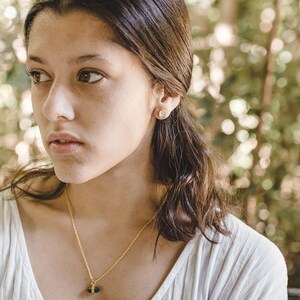 May include: A young woman with long brown hair wearing a white shirt and a gold necklace with a green gemstone pendant. She is wearing a small gold earring in her left ear.