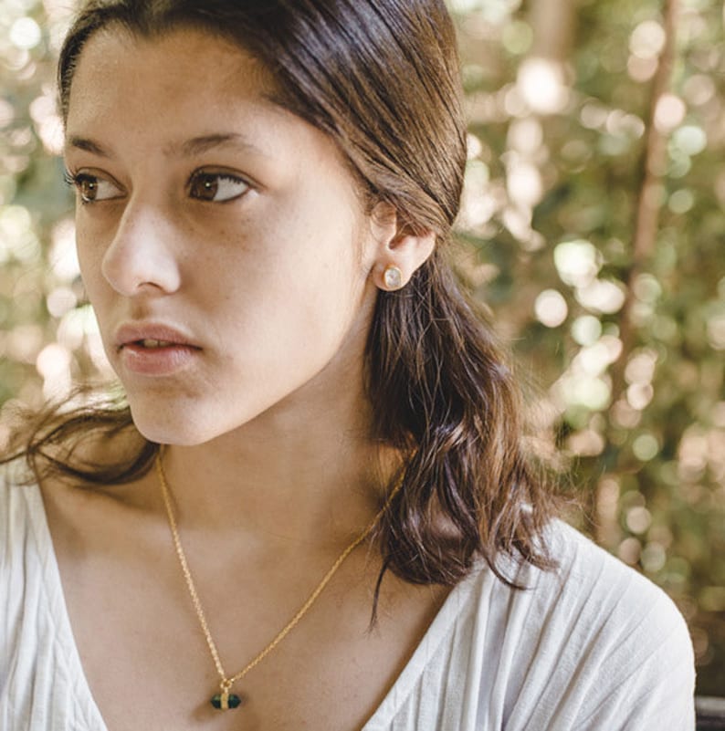 May include: A young woman with long brown hair wearing a white shirt and a gold necklace with a green gemstone pendant. She is wearing a small gold earring in her left ear.