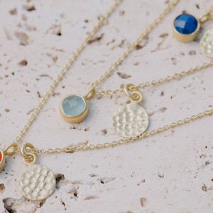 May include: Three gold necklaces with different colored gemstones and hammered gold disc pendants. The necklaces feature a light blue, a dark blue, and an orange gemstone.