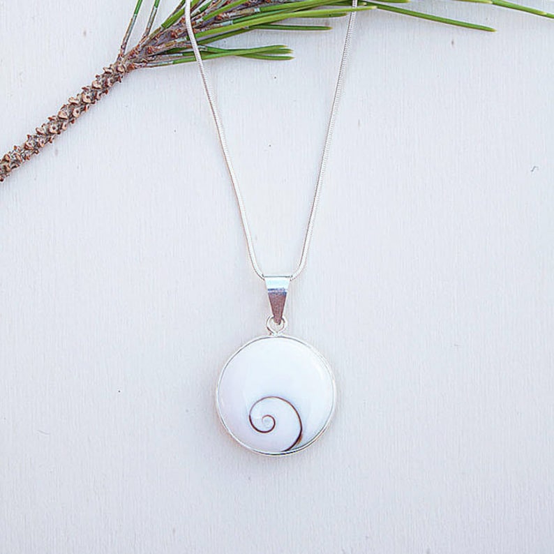 Puede incluir: Un collar de plata con un colgante blanco y redondo con un dise&ntilde;o de espiral marr&oacute;n. El colgante est&aacute; enmarcado en plata y cuelga de una fina cadena de plata. Una ramita de vegetaci&oacute;n est&aacute; en la esquina superior izquierda.