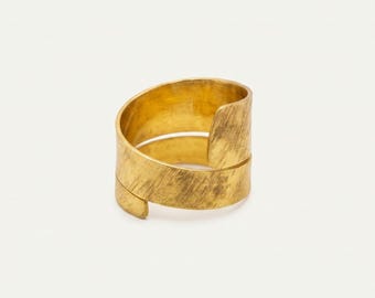 May include: A gold-toned ring featuring a wide, open-ended band design with a textured, brushed finish. The metal surface displays fine, diagonal scratch-like markings across the entire exterior, creating a matte appearance for this circular jewelry piece.