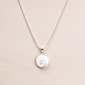 Puede incluir: Un collar de plata con un colgante redondo. El colgante tiene un fondo blanco y un dise&ntilde;o en espiral. El colgante est&aacute; enmarcado en plata y cuelga de una fina cadena de plata. El collar se muestra sobre una superficie de madera clara.