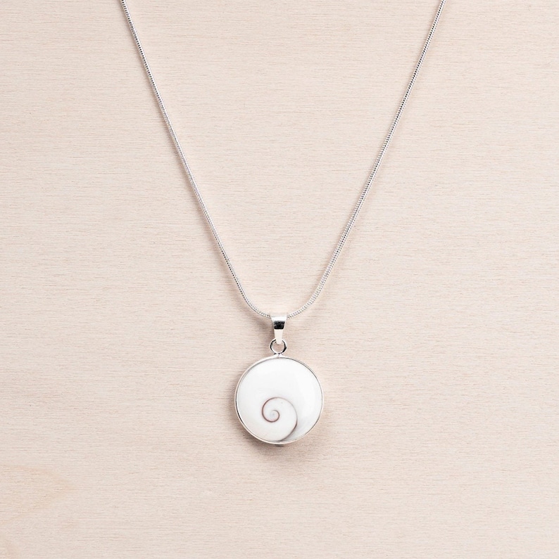 Puede incluir: Un collar de plata con un colgante redondo. El colgante tiene un fondo blanco y un dise&ntilde;o en espiral. El colgante est&aacute; enmarcado en plata y cuelga de una fina cadena de plata. El collar se muestra sobre una superficie de madera clara.