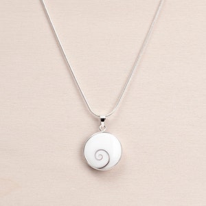 Puede incluir: Un collar plateado con un colgante circular. El colgante presenta un fondo blanco con un dise&ntilde;o en espiral en tonos grises. La cadena del collar es una fina cadena de serpiente plateada.