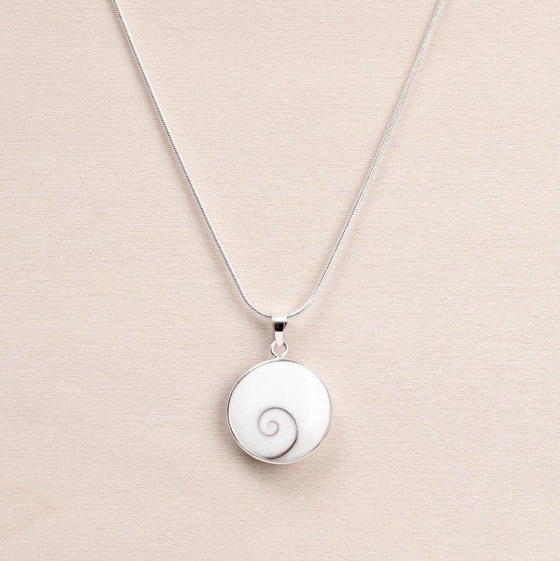 Puede incluir: Un collar plateado con un colgante circular. El colgante presenta un fondo blanco con un dise&ntilde;o en espiral en tonos grises. La cadena del collar es una fina cadena de serpiente plateada.