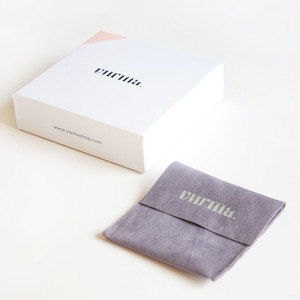 May include: A white box with the word "vurrna." printed on the top and the website address "www.vurmashop.com" printed on the side. The box is sitting on a white surface. A small gray velvet pouch with the word "vurrna." printed on the front is also on the white surface.
