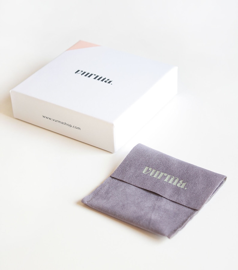 May include: A white box with the word "vurrna." printed on the top and the website address "www.vurmashop.com" printed on the side. The box is sitting on a white surface. A small gray velvet pouch with the word "vurrna." printed on the front is also on the white surface.