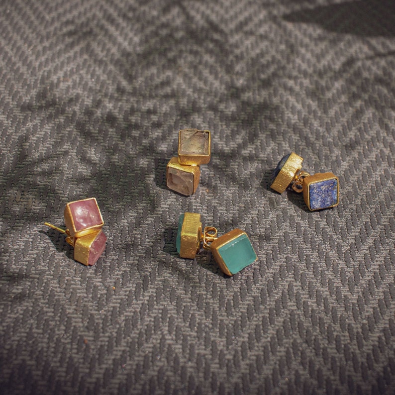 May include: Five pairs of gold earrings with different colored gemstone accents. The earrings feature square-shaped gemstones in shades of pink, blue, green, and clear.