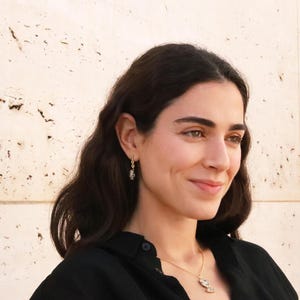 May include: A woman with long dark hair wearing a black shirt and gold jewelry. She is wearing a necklace with a small pendant and earrings with a small charm.