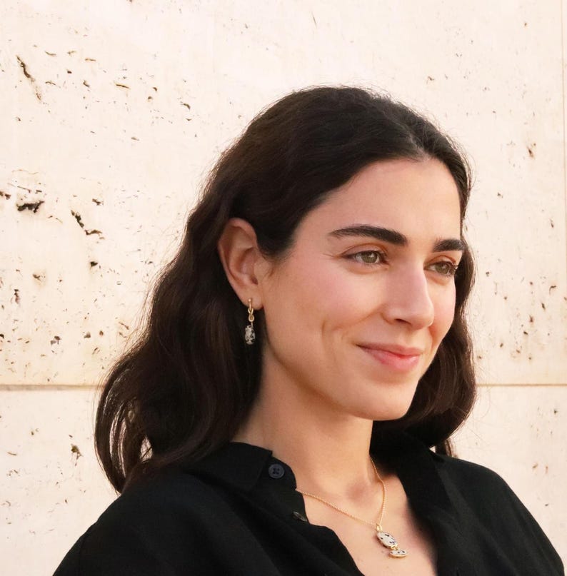 May include: A woman with long dark hair wearing a black shirt and gold jewelry. She is wearing a necklace with a small pendant and earrings with a small charm.