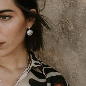 A close-up of a woman with short dark hair wearing elegant silver seashell stud earrings paired with a pearl accent, styled with natural makeup and a patterned outfit, against a soft, breezy background.