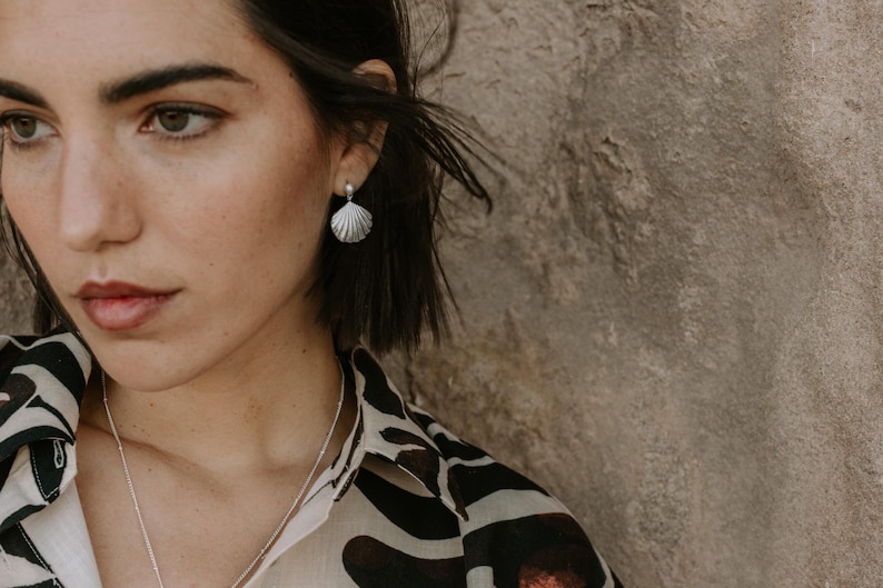 A close-up of a woman with short dark hair wearing elegant silver seashell stud earrings paired with a pearl accent, styled with natural makeup and a patterned outfit, against a soft, breezy background.