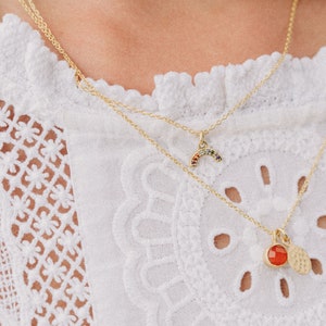 May include: Two gold necklaces with pendants. The top necklace has a rainbow-colored stone pendant shaped like a crescent moon. The bottom necklace has a red stone pendant with a hammered gold disc.