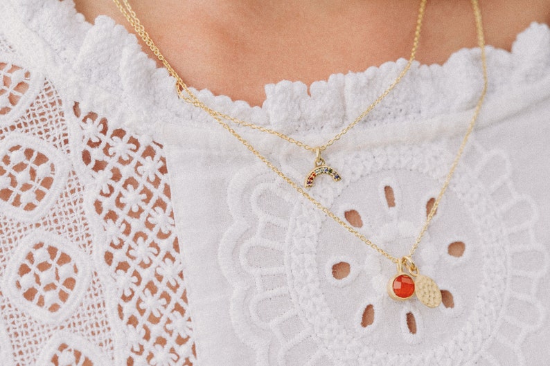 May include: Two gold necklaces with pendants. The top necklace has a rainbow-colored stone pendant shaped like a crescent moon. The bottom necklace has a red stone pendant with a hammered gold disc.