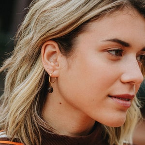 K&ouml;nnte beinhalten: Nahaufnahme eines Frauen Gesichts mit blonden Haaren. Sie tr&auml;gt ein braun-wei&szlig; gestreiftes Shirt und einen goldenen Creolen Ohrring mit einem braunen Edelstein.