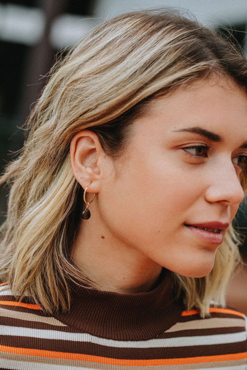 K&ouml;nnte beinhalten: Nahaufnahme eines Frauen Gesichts mit blonden Haaren. Sie tr&auml;gt ein braun-wei&szlig; gestreiftes Shirt und einen goldenen Creolen Ohrring mit einem braunen Edelstein.