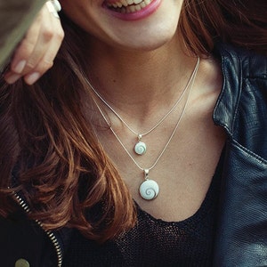 Puede incluir: Dos collares de plata con dise&ntilde;os de colgantes en espiral. Los colgantes son blancos con un patr&oacute;n arremolinado. Los collares est&aacute;n superpuestos, con un colgante m&aacute;s peque&ntilde;o que el otro. El fondo incluye una chaqueta de cuero negra y cabello casta&ntilde;o.