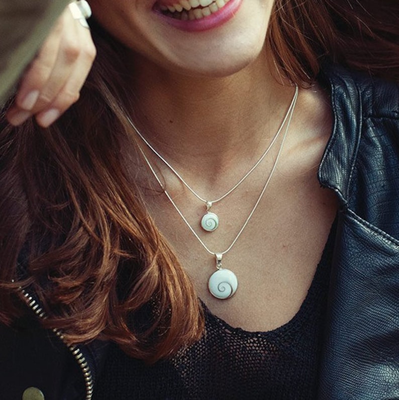 Puede incluir: Dos collares de plata con dise&ntilde;os de colgantes en espiral. Los colgantes son blancos con un patr&oacute;n arremolinado. Los collares est&aacute;n superpuestos, con un colgante m&aacute;s peque&ntilde;o que el otro. El fondo incluye una chaqueta de cuero negra y cabello casta&ntilde;o.