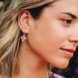 May include: A woman wearing a pink floral dress with a pair of gold earrings featuring three light purple oval-shaped stones.