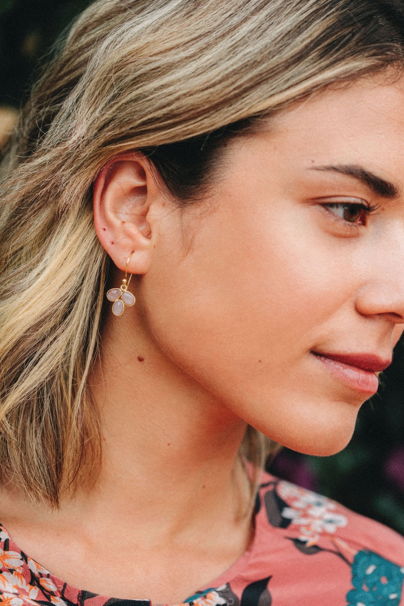 May include: A woman wearing a pink floral dress with a pair of gold earrings featuring three light purple oval-shaped stones.