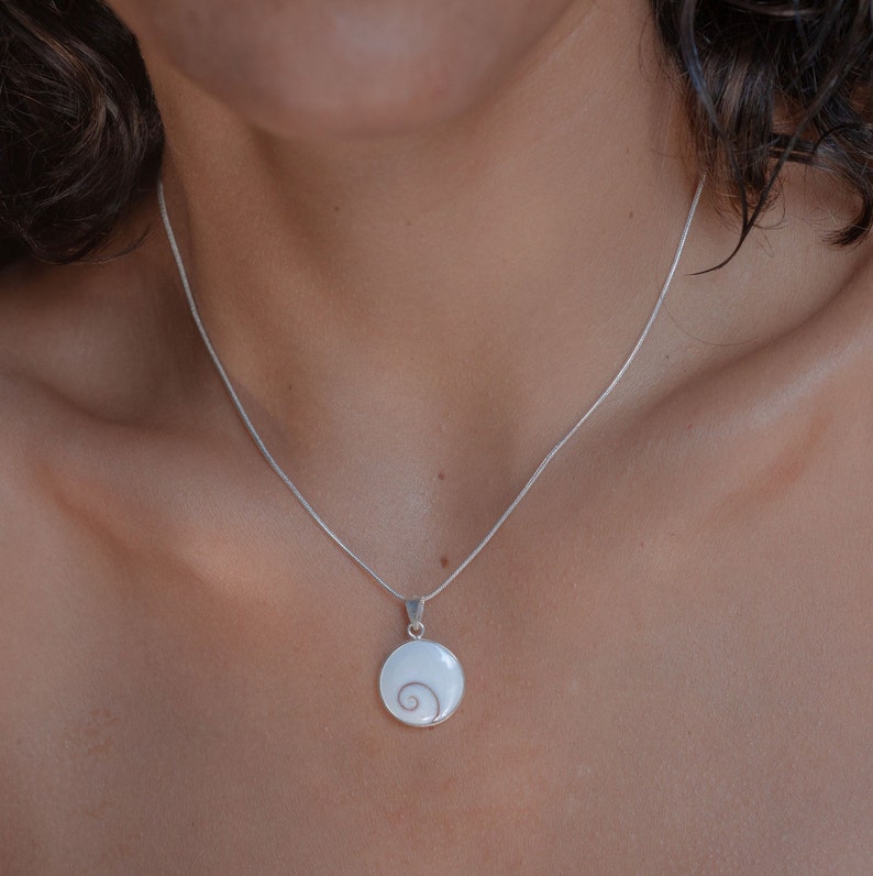 Puede incluir: Un collar de plata con un colgante redondo. El colgante tiene un fondo blanco con un dise&ntilde;o en espiral en el centro. El collar se muestra en el pecho de una persona.
