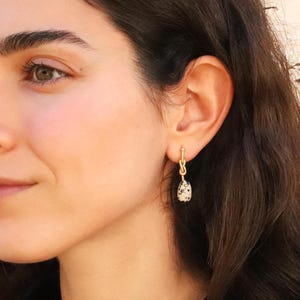 Close-up of a woman wearing a handmade gold brass dangle earring with a Dalmatian jasper gemstone pendant. Elegant and minimalist boho-chic jewelry for women