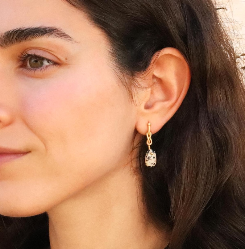 Close-up of a woman wearing a handmade gold brass dangle earring with a Dalmatian jasper gemstone pendant. Elegant and minimalist boho-chic jewelry for women