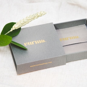 K&ouml;nnte beinhalten: Eine graue Geschenkbox mit einem Schiebedeckel, die Box ist ge&ouml;ffnet und zeigt eine graue Samt-Tasche im Inneren. Die Box hat den Text "Vurma" in Gold auf der Vorderseite und "www.vurmashop.com" in Gold auf der Unterseite gedruckt.