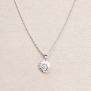 Puede incluir: Un collar de plata con un colgante redondo con un dise&ntilde;o de espiral blanco. El colgante est&aacute; enmarcado en plata y cuelga de una fina cadena de plata. El collar se muestra sobre una superficie de madera clara.