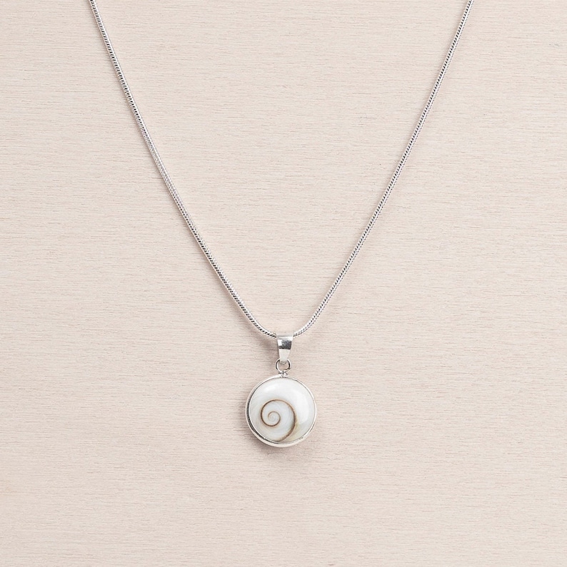 Puede incluir: Un collar de plata con un colgante redondo con un dise&ntilde;o de espiral blanco. El colgante est&aacute; enmarcado en plata y cuelga de una fina cadena de plata. El collar se muestra sobre una superficie de madera clara.