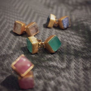 May include: A set of six gold earrings with different colored gemstones. The earrings are square shaped and have a polished gold finish. The gemstones are green, pink, and blue.