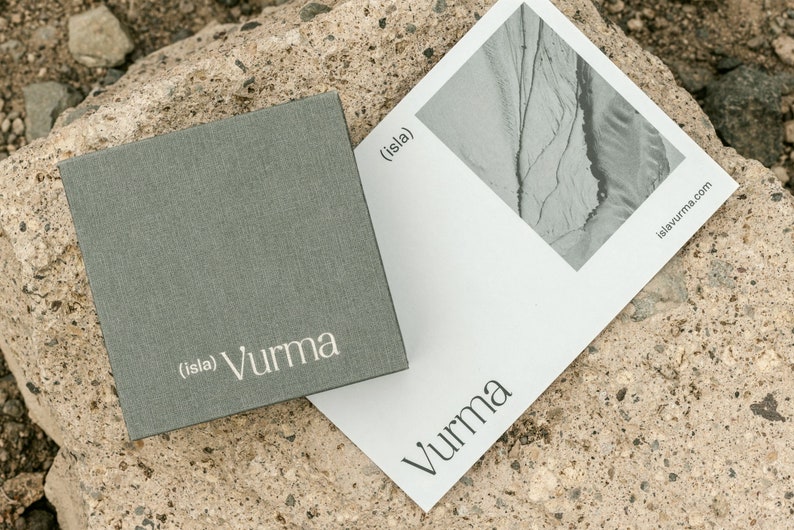May include: A gray box with the text "(isla) Vurma" printed in white. A white card with a black and white photo of a landscape and the text "(isla) Vurma" and "islayvurma.com" printed in black.