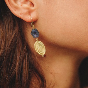 K&ouml;nnte beinhalten: Goldene Ohrringe mit einem blauen Edelstein und einem runden Anh&auml;nger. Der Anh&auml;nger hat ein strukturiertes Design.