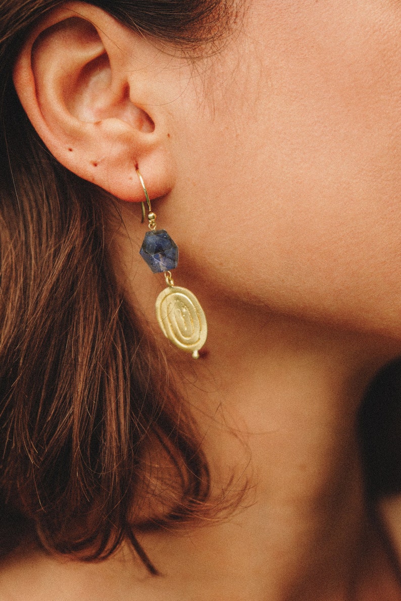 K&ouml;nnte beinhalten: Goldene Ohrringe mit einem blauen Edelstein und einem runden Anh&auml;nger. Der Anh&auml;nger hat ein strukturiertes Design.