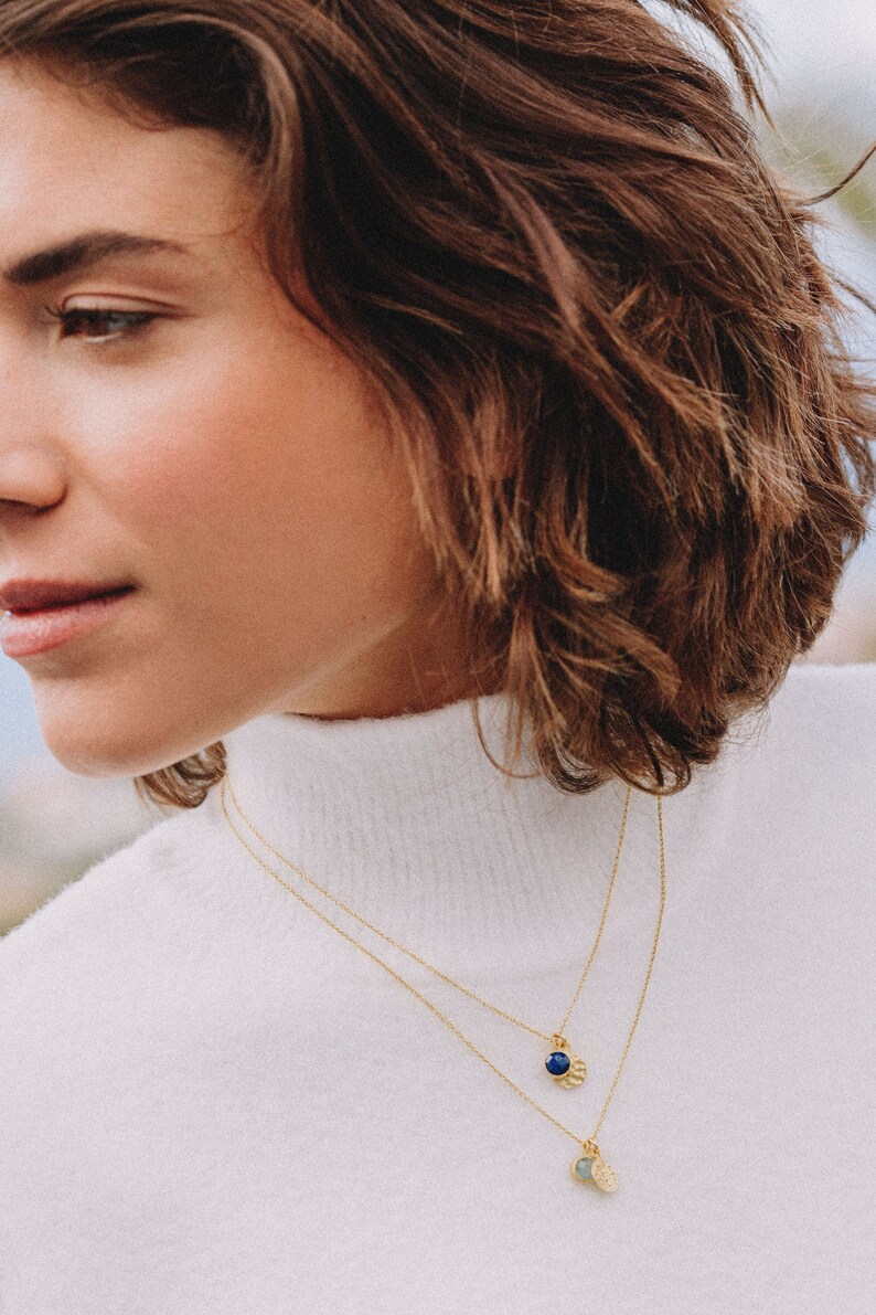 May include: A woman wearing a white turtleneck sweater with a gold chain necklace with two pendants. The pendants are a small round blue stone and a larger round green stone.