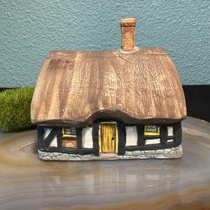 Puede incluir: Una pequeña casa de cerámica con tejado de paja y chimenea de ladrillo. La casa tiene entramado de madera en blanco y negro, una puerta amarilla y pequeñas ventanas. El tejado es marrón y las paredes son blancas y grises.